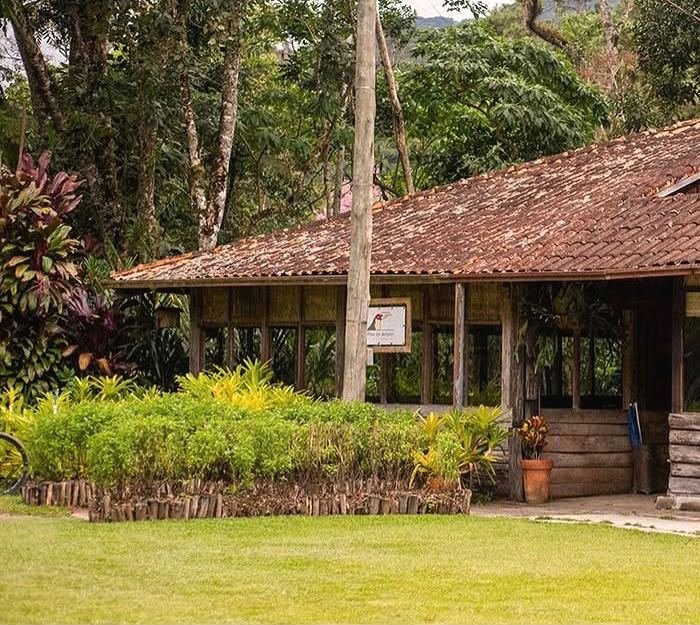 Casa de madeira com telhado de barro do Trilhas do Araçari, cercada por jardim e árvores